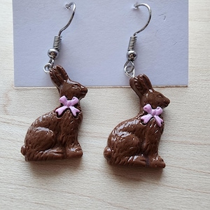 Puede incluir: Un par de pendientes colgantes con un conejito de chocolate marrón con un lazo rosa. Los pendientes están hechos de metal y tienen un cierre de gancho.