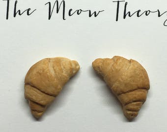 Pode incluir: Um par de brincos em forma de croissant. Os brincos são feitos de um material marrom claro e têm um design de croissant realista.