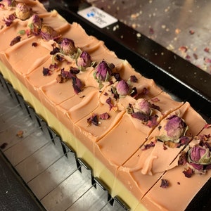 以下が含まれることがあります： 手作りの石鹸の塊で、長方形のバーにカットされています。明るいピーチ色のトップ層、クリーム色のミドル層、濃いピーチ色のボトム層があります。石鹸は、乾燥したバラのつぼみと花びらで飾られています。