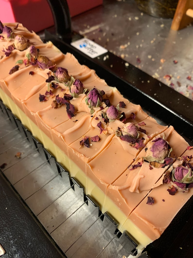 以下が含まれることがあります： 手作りの石鹸の塊で、長方形のバーにカットされています。明るいピーチ色のトップ層、クリーム色のミドル層、濃いピーチ色のボトム層があります。石鹸は、乾燥したバラのつぼみと花びらで飾られています。
