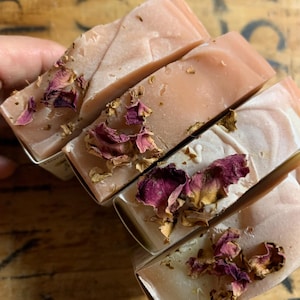 以下が含まれることがあります： 明るいピンク色と乾燥したバラの花びらが上に乗った、手作りの石鹸が4つ。石鹸は長方形で、個別に包装されているようです。石鹸は人の手で持たれています。
