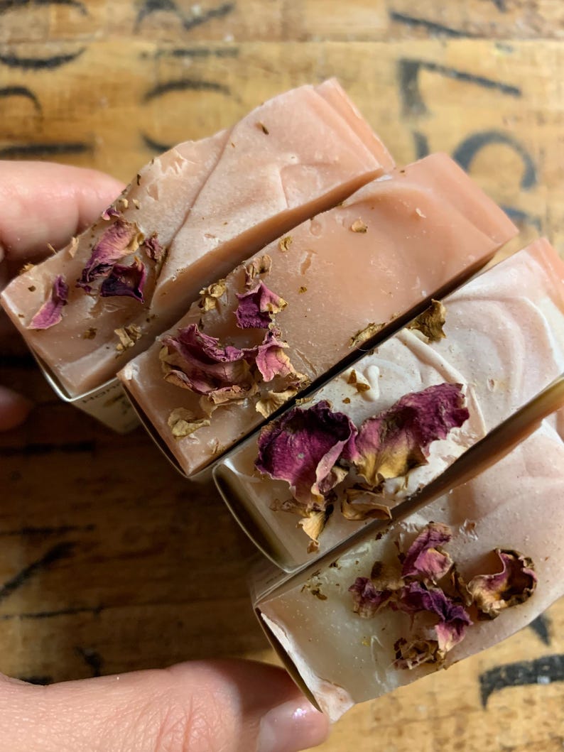 以下が含まれることがあります： 明るいピンク色と乾燥したバラの花びらが上に乗った、手作りの石鹸が4つ。石鹸は長方形で、個別に包装されているようです。石鹸は人の手で持たれています。