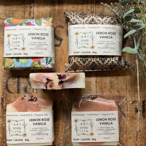 以下が含まれることがあります： 「Lemon Rose Vanilla」と「Art Soap Life」の文字が入った手作りの石鹸の詰め合わせ。石鹸は、模様入りの紙や透明な包装など、さまざまなスタイルで包装されています。石鹸は白とピンクで、ドライフラワーのアクセントが付いています。各石鹸は85gです。