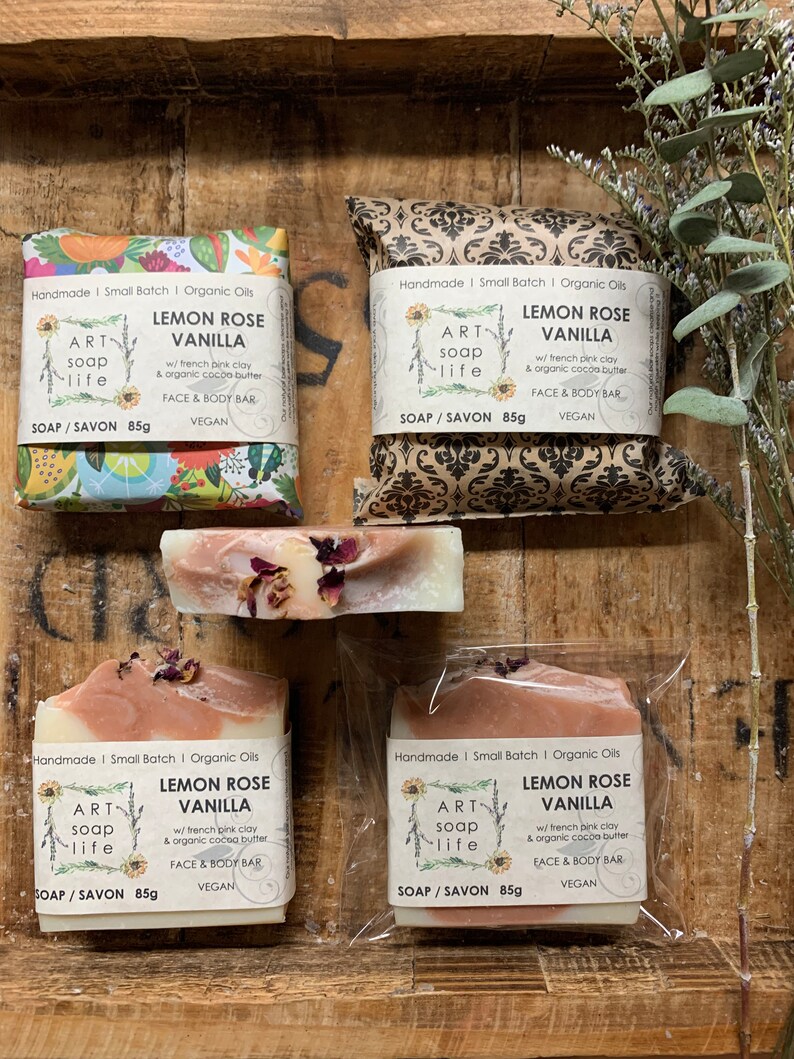 以下が含まれることがあります： 「Lemon Rose Vanilla」と「Art Soap Life」の文字が入った手作りの石鹸の詰め合わせ。石鹸は、模様入りの紙や透明な包装など、さまざまなスタイルで包装されています。石鹸は白とピンクで、ドライフラワーのアクセントが付いています。各石鹸は85gです。