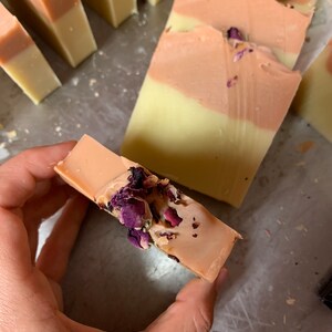 以下が含まれることがあります： 手作りの石鹸バー。明るい黄色のベースとピーチ色のトップレイヤーの2色デザイン。石鹸は乾燥したバラの花びらで飾られています。いくつかのバーが見え、一部は手で持たれています。