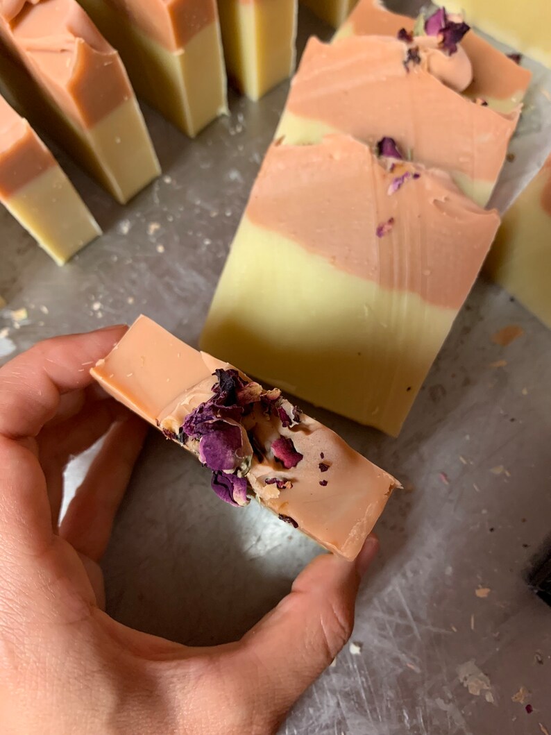 以下が含まれることがあります： 手作りの石鹸バー。明るい黄色のベースとピーチ色のトップレイヤーの2色デザイン。石鹸は乾燥したバラの花びらで飾られています。いくつかのバーが見え、一部は手で持たれています。