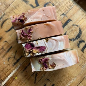 以下が含まれることがあります： 手作りの石鹸が4つ積み重ねられており、薄いピンクと白の大理石模様が特徴です。各石鹸には乾燥したバラの花びらが飾られています。石鹸は素朴な木製の表面に配置されており、自然な美しさを醸し出しています。