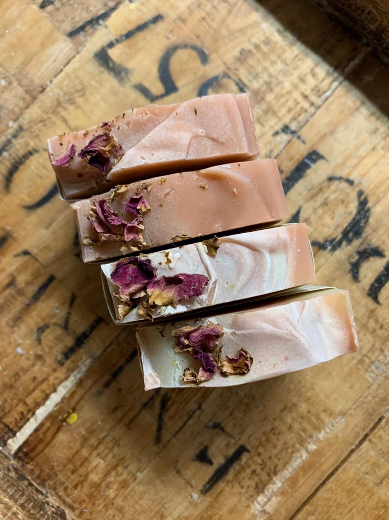以下が含まれることがあります： 手作りの石鹸が4つ積み重ねられており、薄いピンクと白の大理石模様が特徴です。各石鹸には乾燥したバラの花びらが飾られています。石鹸は素朴な木製の表面に配置されており、自然な美しさを醸し出しています。