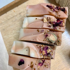 以下が含まれることがあります： 木製のトレイに並べられた、手作りの石鹸6個。石鹸はピンクと白の渦巻き模様で、乾燥したバラの花びらとつぼみが飾られています。長方形の石鹸は、切りたてのように見えます。