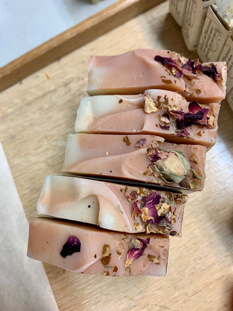 以下が含まれることがあります： 木製のトレイに並べられた、手作りの石鹸6個。石鹸はピンクと白の渦巻き模様で、乾燥したバラの花びらとつぼみが飾られています。長方形の石鹸は、切りたてのように見えます。