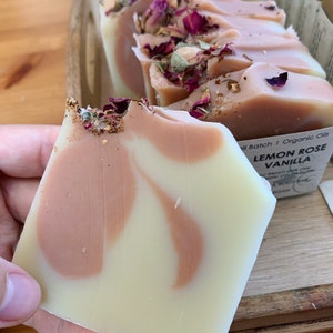 以下が含まれることがあります： クリームとピーチ色の渦巻き模様の手作り石鹸バー。乾燥バラの花びらと金箔で飾られています。「Lemon Rose Vanilla」と「Organic Oils」と書かれたラベルが付いています。