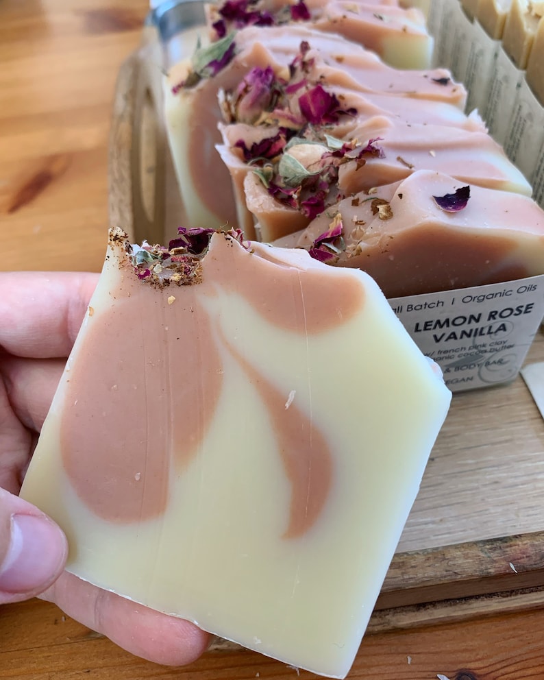 以下が含まれることがあります： クリームとピーチ色の渦巻き模様の手作り石鹸バー。乾燥バラの花びらと金箔で飾られています。「Lemon Rose Vanilla」と「Organic Oils」と書かれたラベルが付いています。