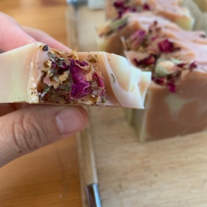 以下が含まれることがあります： 手作りの石鹸のクローズアップ。薄いピンクと白の渦巻き模様。石鹸は乾燥したバラの花びらとハーブで飾られています。背景にはいくつかの石鹸が見え、自然なスキンケア製品を示唆しています。