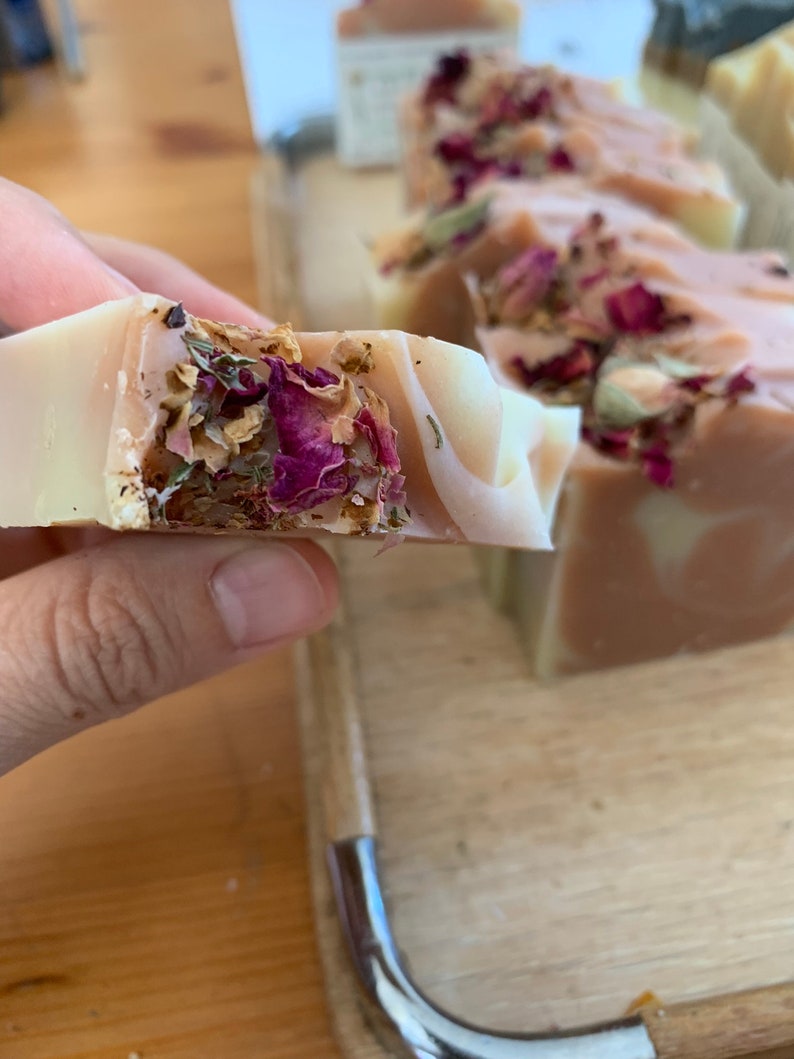 以下が含まれることがあります： 手作りの石鹸のクローズアップ。薄いピンクと白の渦巻き模様。石鹸は乾燥したバラの花びらとハーブで飾られています。背景にはいくつかの石鹸が見え、自然なスキンケア製品を示唆しています。