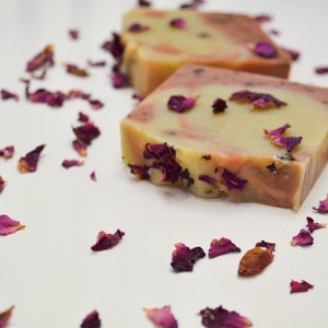 以下が含まれることがあります： 手作りの石鹸2つ。薄い黄色とピンクの渦巻き模様で、乾燥したバラの花びらで飾られています。長方形の石鹸は白い表面に置かれ、周りにバラの花びらが散らばっています。
