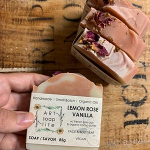 以下が含まれることがあります： Art Soap Lifeのレモンローズバニラフェイス＆ボディバー。石鹸はピンクと白で、乾燥したバラの花びらがトッピングされています。ラベルには「Handmade | Small Batch | Organic Oils」と「Vegan」と書かれています。石鹸の重量は85gです。