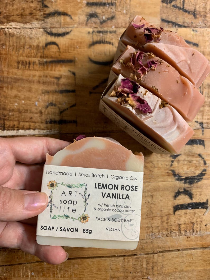 以下が含まれることがあります： Art Soap Lifeのレモンローズバニラフェイス＆ボディバー。石鹸はピンクと白で、乾燥したバラの花びらがトッピングされています。ラベルには「Handmade | Small Batch | Organic Oils」と「Vegan」と書かれています。石鹸の重量は85gです。