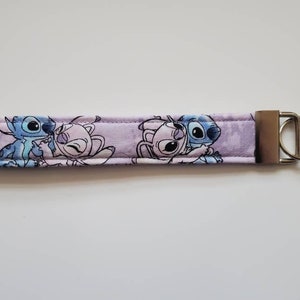 Op de afbeelding: Een paarse sleutelhanger van stof met een blauw en roze cartoonfiguurontwerp. De sleutelhanger heeft een zilveren metalen ring.