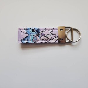 Op de afbeelding: Een paarse stoffen sleutelhanger met een cartoon afbeelding van Stitch en Angel uit de Disney-film Lilo & Stitch.
