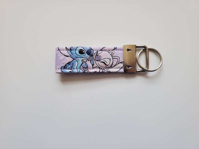 Op de afbeelding: Een paarse stoffen sleutelhanger met een cartoon afbeelding van Stitch en Angel uit de Disney-film Lilo & Stitch.