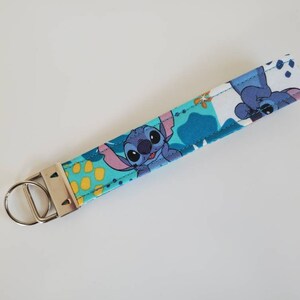Op de afbeelding: Een blauwe en witte stoffen sleutelhanger met een cartoonfiguur van Stitch uit Lilo & Stitch. De sleutelhanger heeft een zilveren metalen sleutelring.