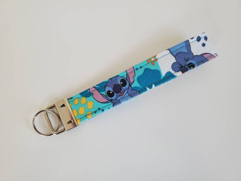 Op de afbeelding: Een blauwe en witte stoffen sleutelhanger met een cartoonfiguur van Stitch uit Lilo & Stitch. De sleutelhanger heeft een zilveren metalen sleutelring.