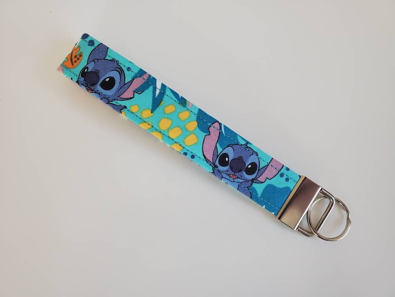 Op de afbeelding: Een blauwe en groene sleutelhanger met een cartoonfiguur van Stitch uit de Disney-film Lilo & Stitch. De sleutelhanger heeft een zilveren metalen ring.