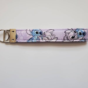 Op de afbeelding: Een paarse sleutelhanger van stof met een zilveren sleutelring. De sleutelhanger heeft een cartoon afbeelding van Stitch en Angel uit de Disney-film Lilo & Stitch.