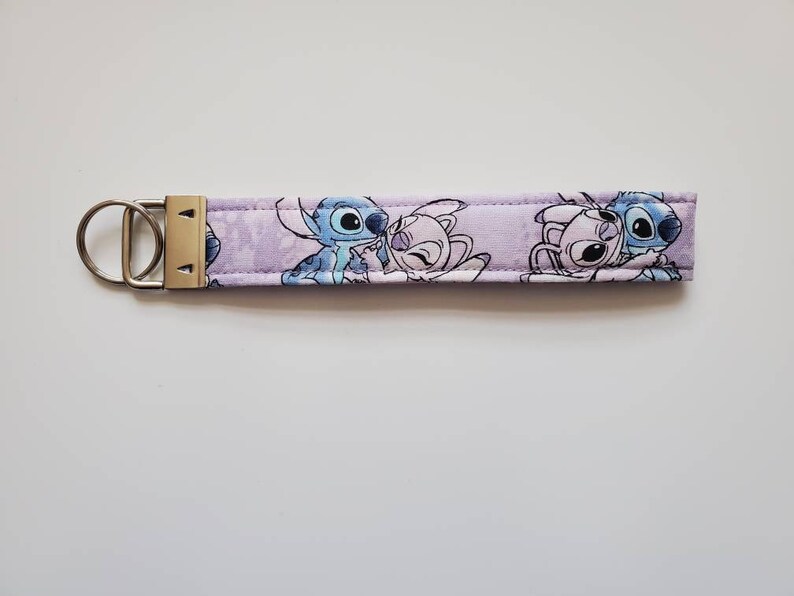 Op de afbeelding: Een paarse sleutelhanger van stof met een zilveren sleutelring. De sleutelhanger heeft een cartoon afbeelding van Stitch en Angel uit de Disney-film Lilo & Stitch.