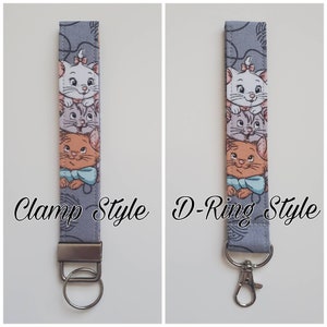 Op de afbeelding: Een grijze sleutelhanger van stof met een ontwerp van drie cartoonkatten. De sleutelhanger wordt in twee stijlen getoond: klem stijl en D-ring stijl.
