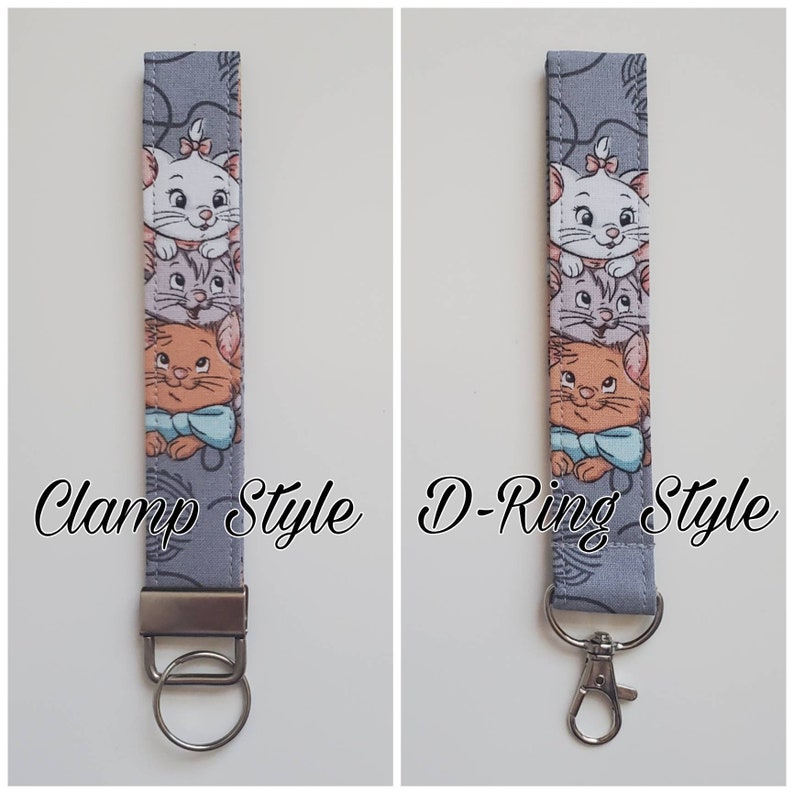 Op de afbeelding: Een grijze sleutelhanger van stof met een ontwerp van drie cartoonkatten. De sleutelhanger wordt in twee stijlen getoond: klem stijl en D-ring stijl.