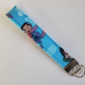 Op de afbeelding: Een blauwe en witte sleutelhanger met een patroon van Stitch en Lilo uit de Disney-film Lilo & Stitch. De sleutelhanger heeft een zilveren sleutelring.