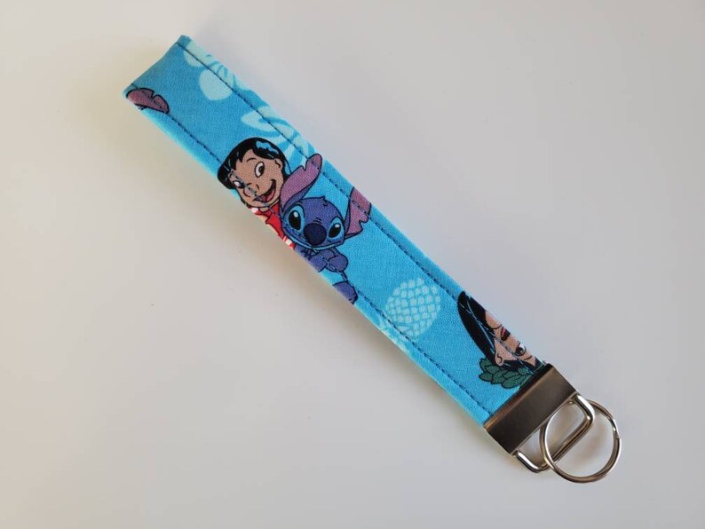 Op de afbeelding: Een blauwe en witte sleutelhanger met een patroon van Stitch en Lilo uit de Disney-film Lilo & Stitch. De sleutelhanger heeft een zilveren sleutelring.