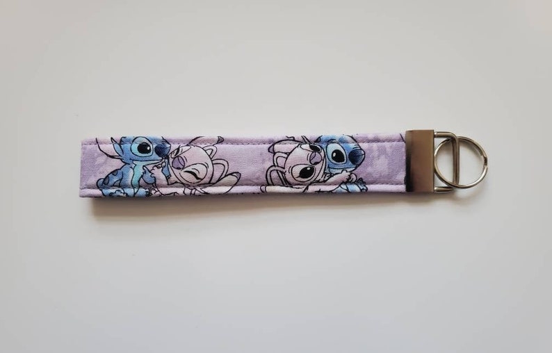 Op de afbeelding: Een paarse sleutelhanger van stof met een blauw en roze cartoonfiguurontwerp. De sleutelhanger heeft een zilveren metalen ring.