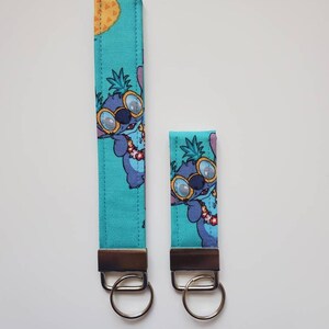 Op de afbeelding: Twee blauwe en groene sleutelhangers met een cartoonfiguur dat een zonnebril en een lei draagt. Het personage bevindt zich op een turquoise achtergrond met ananasontwerpen. De sleutelhangers zijn bevestigd aan zilveren sleutelringen.