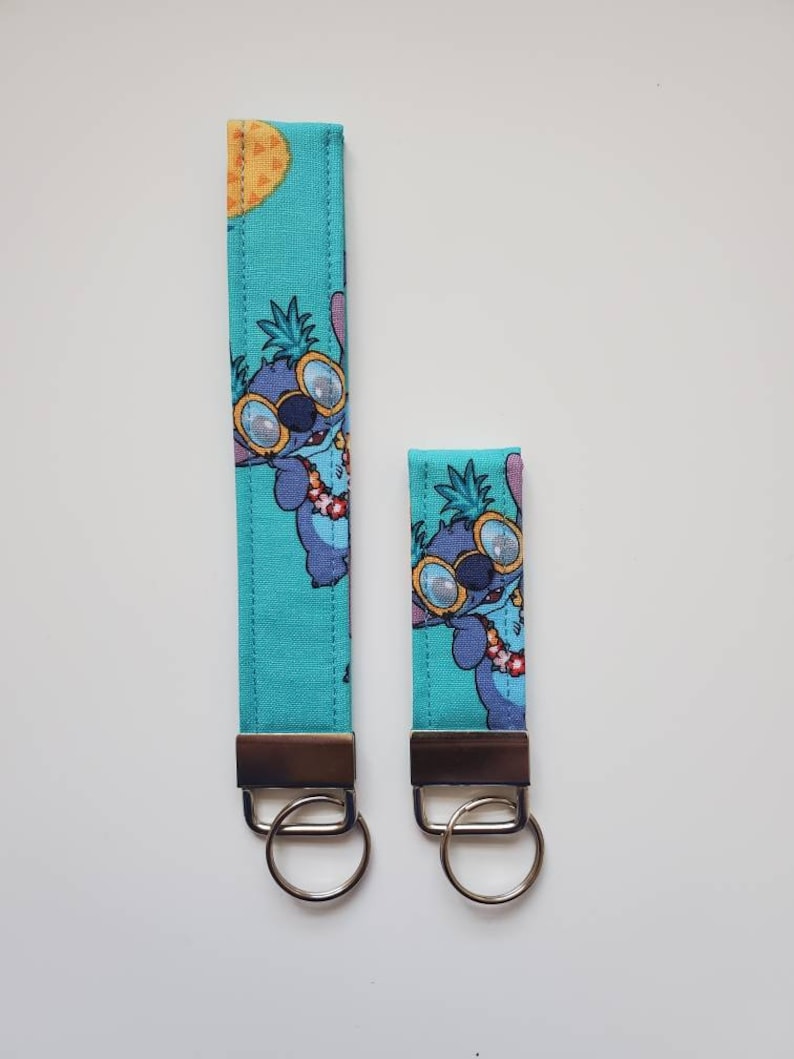 Op de afbeelding: Twee blauwe en groene sleutelhangers met een cartoonfiguur dat een zonnebril en een lei draagt. Het personage bevindt zich op een turquoise achtergrond met ananasontwerpen. De sleutelhangers zijn bevestigd aan zilveren sleutelringen.