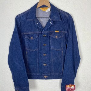 May include: A dark blue denim jacket with a yellow "RUSTLER" label on the pocket and a red "RUSTLER" tag on the sleeve. The jacket has a button closure and two front pockets.