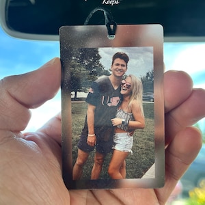 May include: A rectangular car air freshener featuring a printed photograph of a couple standing outdoors. The man wears a dark t-shirt and shorts, while the woman wears a striped top and denim shorts. The item has a black string for hanging.
