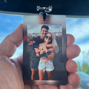 May include: A photo car air freshener with a picture of a couple hugging in front of a bridge. The text "Memorable Keeps" is printed on the top of the air freshener.