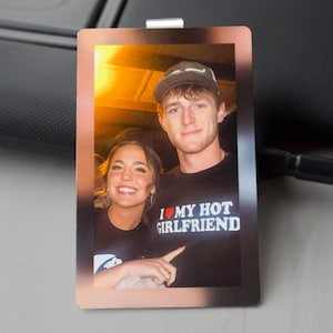 May include: A rectangular photo with a copper-colored border. The photo features a smiling couple. The man wears a black t-shirt with the text "I ❤️ MY HOT GIRLFRIEND" in white and red. The woman is also wearing a black t-shirt.