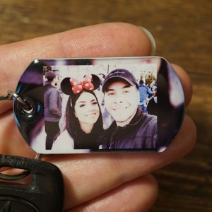 以下が含まれることがあります： カップルが写っている銀色のキーホルダー。女性は赤と黒のミッキーマウスの耳をつけています。男性は黒い野球帽をかぶっています。写真は銀色のタグに印刷されています。