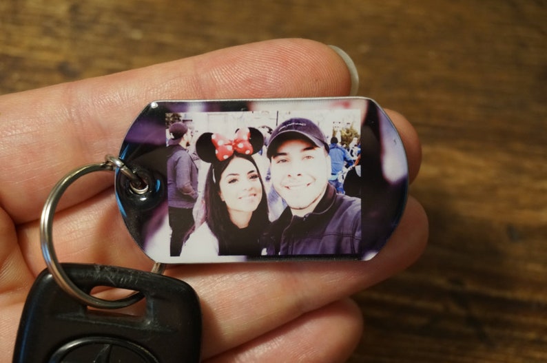 以下が含まれることがあります： カップルが写っている銀色のキーホルダー。女性は赤と黒のミッキーマウスの耳をつけています。男性は黒い野球帽をかぶっています。写真は銀色のタグに印刷されています。