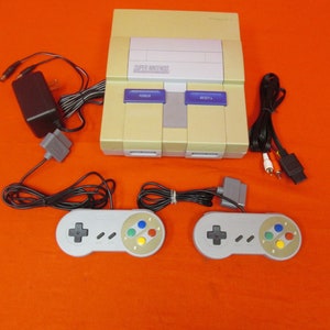 May include: A Super Nintendo Entertainment System (SNES) console in beige with two gray controllers, a power adapter, and AV cables. The console has the text "SUPER NINTENDO" printed on the front.