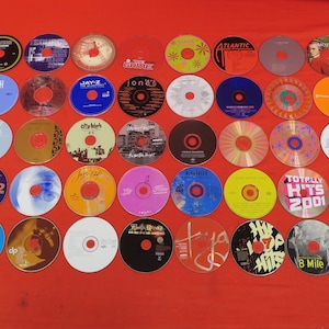 May include: A collection of 40 compact discs (CDs) with various titles and artists. The CDs are arranged in a grid pattern on a red surface. Some of the titles include "Cash", "Naughty by Nature", "Jay-Z", "Jon", "Atlantic", "Cuba LA", "Totally Hits 2001", "8 Mile", and "2".
