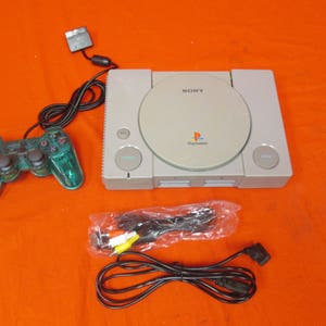 May include: A vintage PlayStation console with a transparent green controller, power cord, and AV cables. The console is light gray with the Sony and PlayStation logos. The controller has black buttons and a directional pad.