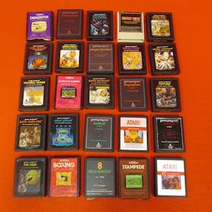 May include: A collection of 20 vintage Atari video game cartridges, including titles like "Dragster", "Warlords", "Haunted House", "Super Breakout", "Pac-Man", and "Boxing". The cartridges are arranged in four rows of five, on a bright orange surface.