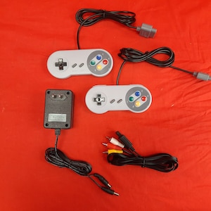 Puede incluir: Un conjunto de consola de juegos retro con dos mandos grises, un adaptador de corriente negro y un cable con conectores rojo, amarillo y blanco. El conjunto está sobre una superficie roja.