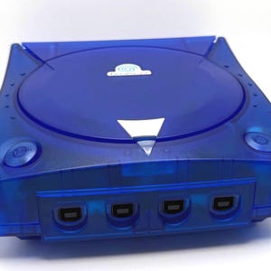 Könnte beinhalten: Eine durchsichtige blaue Sega Dreamcast Videospielkonsole mit einem runden Deckel für das Laufwerk, auf dem das Dreamcast-Logo zu sehen ist, einem silbernen dreieckigen Öffnungsknopf und vier Controller-Anschlüssen an der Vorderseite.