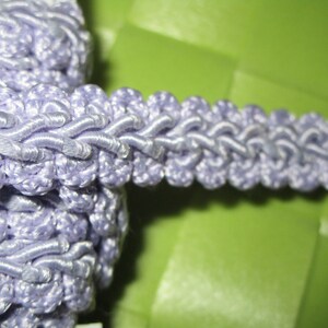 May include: A close-up of a lavender-colored braided trim with a textured, rope-like design. The trim is made of a soft, pliable material and has a delicate, feminine look.