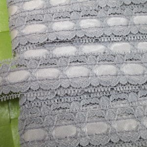 May include: Grey lace trim with a scalloped edge and a repeating pattern of oval shapes. The lace is delicate and has a soft, feminine feel.